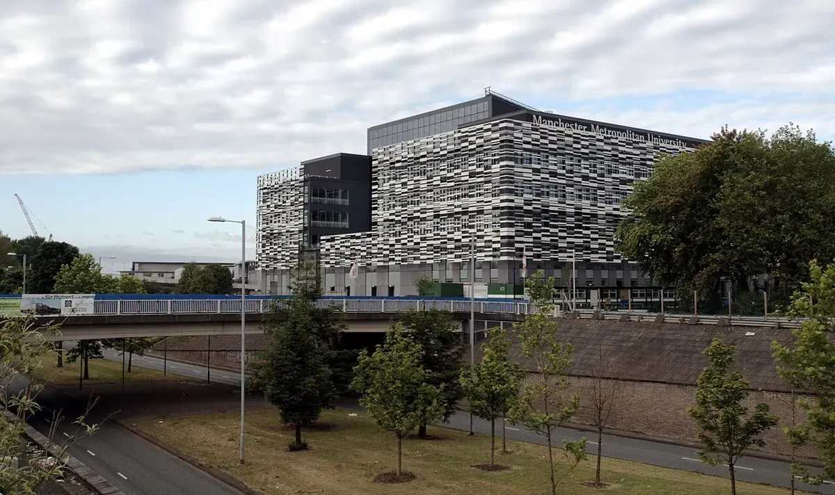 Модерна сграда на Manchester Metropolitan University с черно-бяла фасада, мост, път и млади дървета отпред
